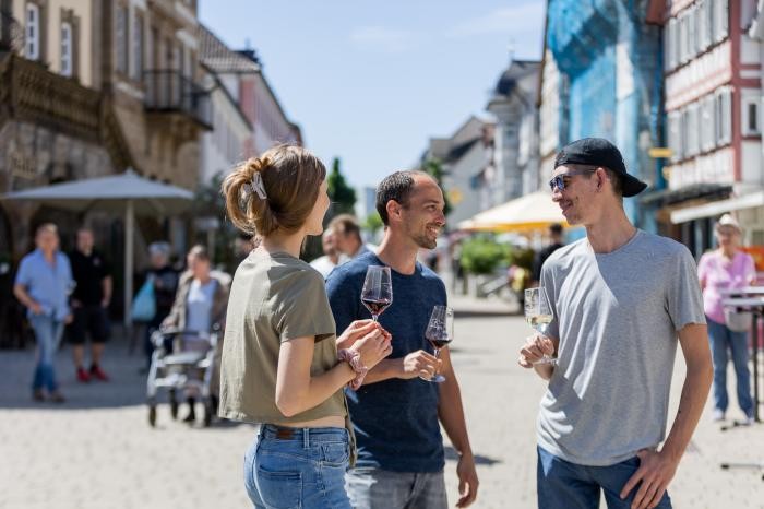 Drei junge Leute stehen in der Fußgängerzone mit Weingläsern in der Hand. 