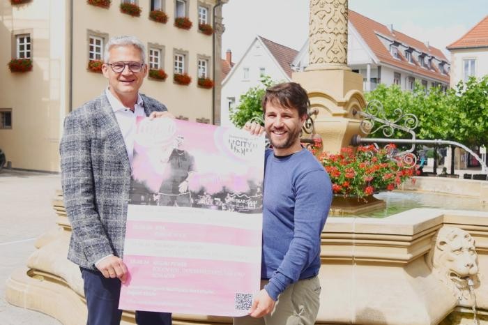 OB Hertwig und Amtsleiter Kiefer stehen vor dem Marktplatzbrunnen und halten das Plakat in die Kamera.  