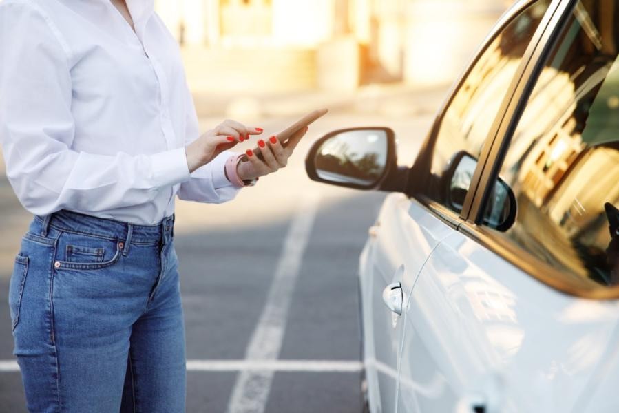 Eine Frau steht vor einem Auto und hält ein Smartphone in der Hand.  