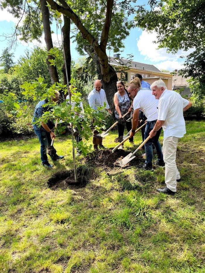 Acht Personen stehen im Kreis und haben Schaufeln in der Hand. Sie pflanzen einen Baum.