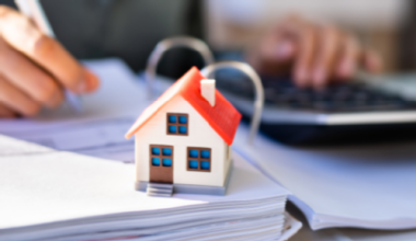 Foto: Adobe Stock Ein Modellhaus steht auf einem Ringbuchordner. Im Hintergrund macht die Hand einer Person Noitzen in diesen Ordner.