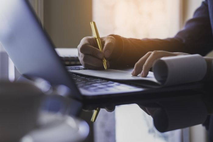 Eine Person hält einen Stift und macht sich Notizen auf einem Papier. Davor steht ein Laptop.