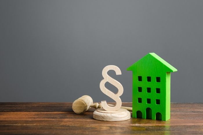 Foto: Adobe Stock Ein grünes Modell eines Hauses steht neben einem hölzernen Paragraphen-Symbol.